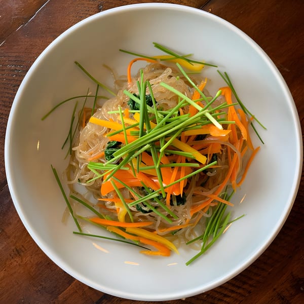 A white bowl of Korean Japchae noodles with carrots, bell peppers, and chives on a wooden table.