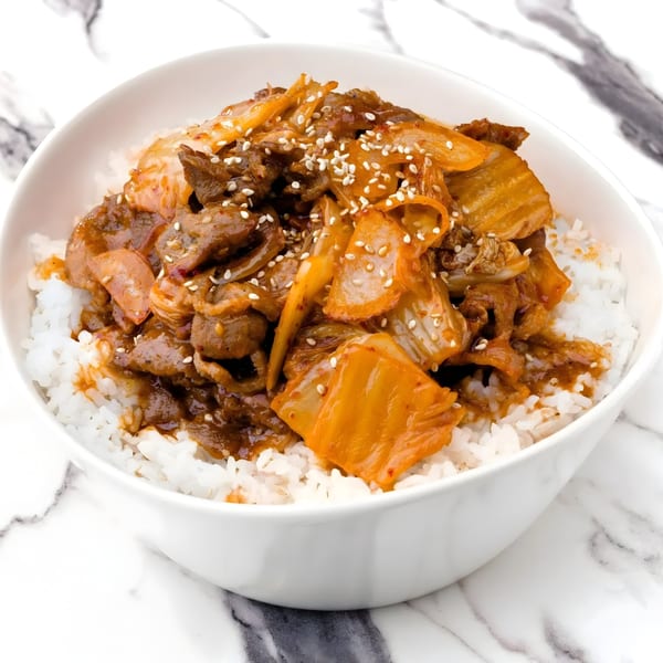 A white bowl filled with white rice, topped with Korean kimchi and beef, sprinkled with sesame seeds.